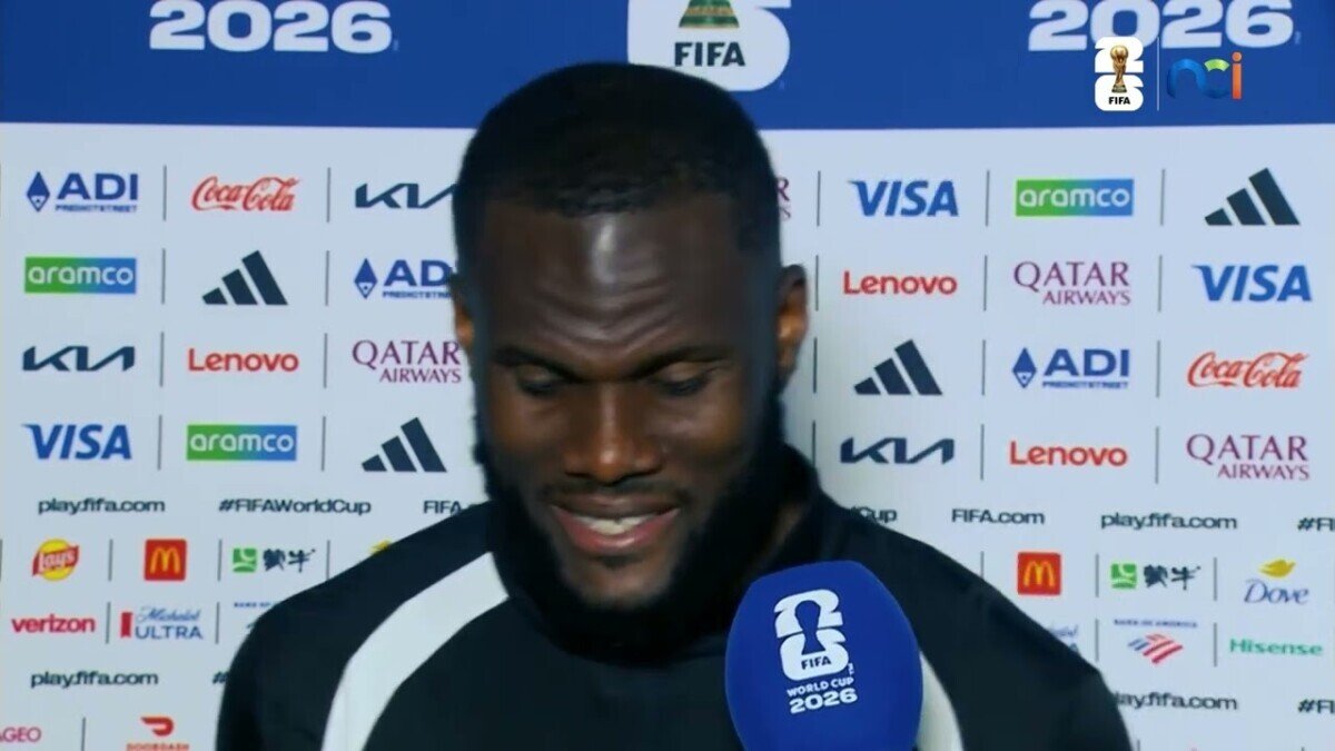 Male footballer being interviewed at a FIFA press conference, holding a blue World Cup microphone with sponsor logos on the backdrop behind him.